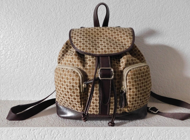 May include: A brown and tan patterned backpack with a brown leather trim and a silver buckle. The backpack has two front pockets and a drawstring closure.