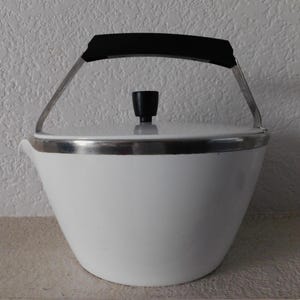 May include: A white, vintage tea kettle with a black handle and lid knob. The kettle has a silver rim and a curved spout. The handle is made of metal and plastic. The kettle is sitting on a light-colored surface.