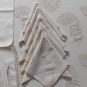 Pu&ograve; includere: Tovaglioli di lino color crema con un delicato design ricamato, impilati ordinatamente su una tovaglia abbinata. I tovaglioli presentano un sottile motivo a foglie, che completa il motivo a foglie pi&ugrave; grande sulla tovaglia. Un set di eleganti tovaglie.