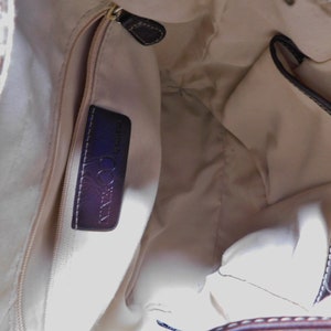 May include: The inside of a beige handbag with a brown leather trim. The bag has a zippered pocket with a black leather label that reads "COCCINELLA" in white lettering.