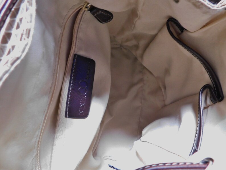 May include: The inside of a beige handbag with a brown leather trim. The bag has a zippered pocket with a black leather label that reads "COCCINELLA" in white lettering.