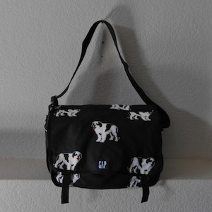 May include: Black messenger bag with a white and black dog print. The bag has a black adjustable strap and a black zipper closure. The bag has the word "GAP" printed on the front.