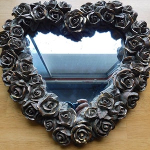 May include: A heart-shaped mirror with a decorative frame made of sculpted roses. The frame is a dark brown color and the mirror is clear.