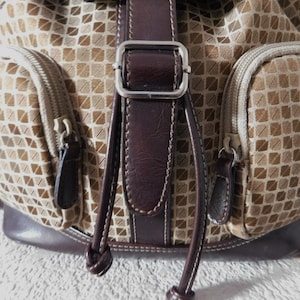 May include: A brown and beige patterned backpack with a brown leather strap and buckle. The backpack has two zippered pockets on the front.
