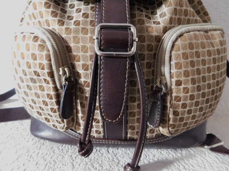 May include: A brown and beige patterned backpack with a brown leather strap and buckle. The backpack has two zippered pockets on the front.
