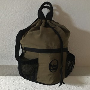 May include: A green drawstring bag with black straps and a zippered pocket. The bag has a mesh pocket on each side and a black logo with a tree and a circle on the front.