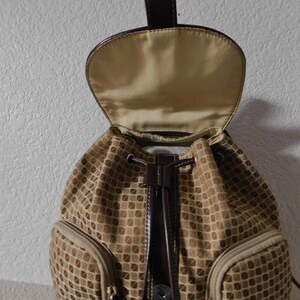 May include: A brown and tan patterned drawstring backpack with a brown leather flap and a brown leather drawstring. The backpack has two zippered pockets on the front.