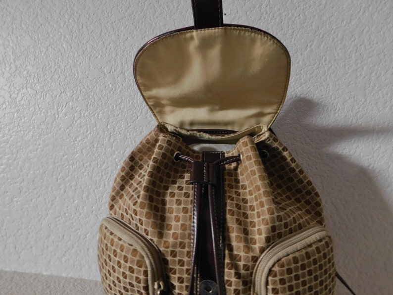 May include: A brown and tan patterned drawstring backpack with a brown leather flap and a brown leather drawstring. The backpack has two zippered pockets on the front.