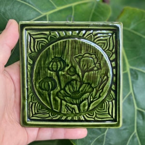 May include: A square, ceramic wall tile with a glossy, green finish. The tile features a raised floral design within a circular frame, surrounded by an ornate border. The flowers include poppy buds and blooms. The tile is held by a hand.