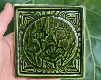 May include: A square, ceramic wall tile with a glossy, green finish. The tile features a raised floral design within a circular frame, surrounded by an ornate border. The flowers include poppy buds and blooms. The tile is held by a hand.