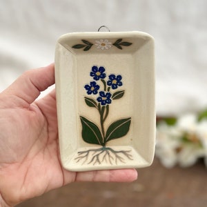 May include: Cream-colored ceramic wall hanging with a floral design. The rectangular piece features a central image of blue flowers with green leaves and a root system. A small daisy and green leaves are at the top. A metal loop is attached for hanging.