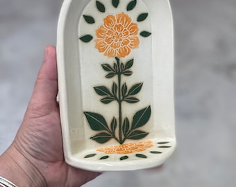 May include: Cream-colored ceramic soap dish with an arched top. The dish features a central orange flower design with green leaves and accents. The soap dish is rectangular with a curved top and a small ledge at the bottom.