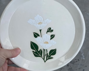 Puede incluir: Un plato de cerámica color crema con un borde elevado. El plato presenta un diseño floral blanco con hojas verdes y un centro amarillo. El plato es redondo y parece hecho a mano.