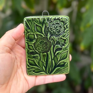 May include: A rectangular ceramic wall plaque with a glossy dark green glaze featuring a relief-carved floral design of two blooming flowers surrounded by leaves and stems. The plaque includes a small metal wire loop at the top for hanging.