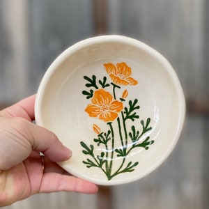 May include: A small, off-white ceramic bowl with a hand-painted floral design. The flowers are orange, with green stems and leaves. The bowl is round and has a slightly uneven rim, suggesting it is handmade.