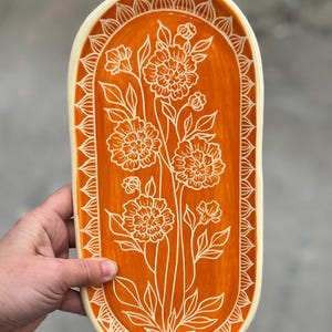 May include: An orange ceramic serving tray with a white floral design. The oval-shaped tray features a detailed white floral pattern in the center and a decorative border. The tray is held by a person.