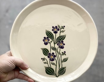 May include: A white ceramic plate with a floral design. The flowers are purple with yellow centers and green leaves.