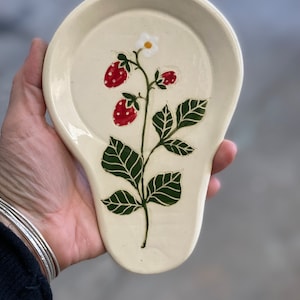 May include: Cream-colored ceramic spoon rest with a hand-painted strawberry plant design. The design features red strawberries with white dots, green leaves, and a small white flower. The spoon rest is oval-shaped with a curved handle.