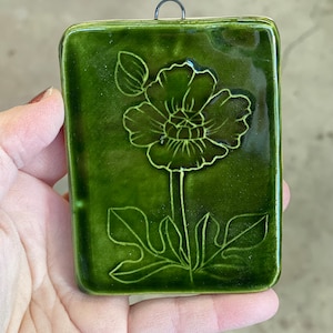 May include: A rectangular, glossy, dark green ceramic tile with a raised floral design. The flower and leaves are outlined in a lighter green. A metal loop is attached for hanging. The tile is held in a hand.