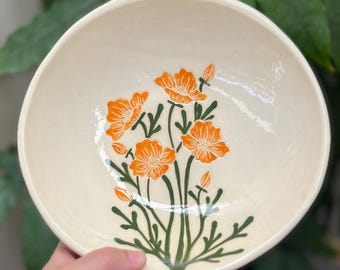 Puede incluir: Cuenco de cerámica color crema con un diseño floral naranja y tallos y hojas verdes. El cuenco tiene forma redonda y un borde irregular. El diseño pintado a mano le da un aspecto artesanal único. Ideal para servir alimentos.