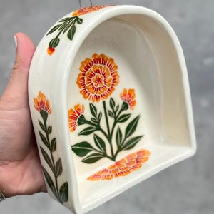 May include: Cream-colored ceramic wall pocket with an arched top and a floral design. The flowers are orange and the leaves are green. A metal wire is attached for hanging.