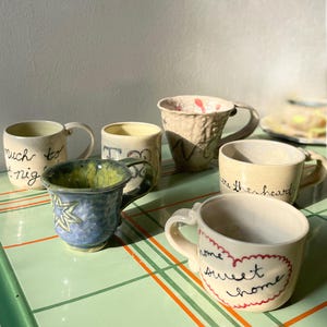 May include: A collection of six ceramic mugs with various finishes and designs. One mug features the text "home sweet home" inside a red heart, another says "much to night", and a third says "where the heart". Some mugs have textured surfaces, while others are smooth with blue, yellow, and cream glazes.