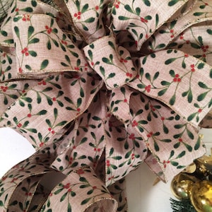 K&ouml;nnte beinhalten: Eine gro&szlig;e, festliche Schleife aus Burlap-Band mit einem Muster aus gr&uuml;nen und roten Stechpalmenbeeren. Das Band hat einen goldenen Glitzerbesatz.