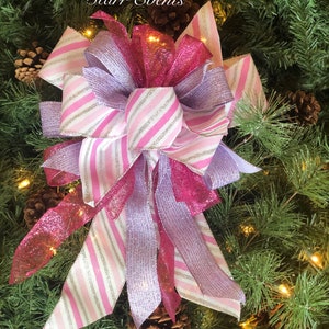K&ouml;nnte beinhalten: Eine rosa-wei&szlig;e gestreifte Schleife mit silbernen Glitzerakzenten, gebunden mit einem rosa-violetten glitzernden Band. Die Schleife befindet sich auf einem gr&uuml;nen Weihnachtsbaum.
