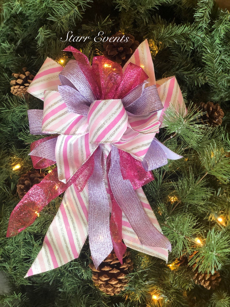 K&ouml;nnte beinhalten: Eine rosa-wei&szlig;e gestreifte Schleife mit silbernen Glitzerakzenten, gebunden mit einem rosa-violetten glitzernden Band. Die Schleife befindet sich auf einem gr&uuml;nen Weihnachtsbaum.