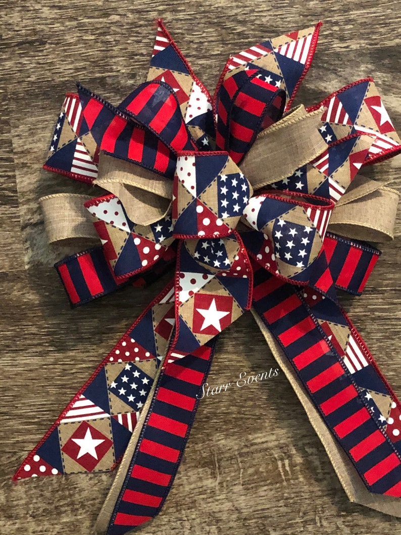 May include: A red, white, and blue patriotic bow with a burlap ribbon and a patchwork design featuring stars and polka dots. The bow is tied with a knot and has two long tails. The text "Starry Events" is visible on the bow.