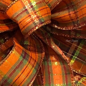 May include: A close-up of a large, orange, green, and gold plaid bow with a shimmery finish. The bow is made of ribbon and is tied in a knot. The bow is perfect for adding a touch of fall decor to any space.