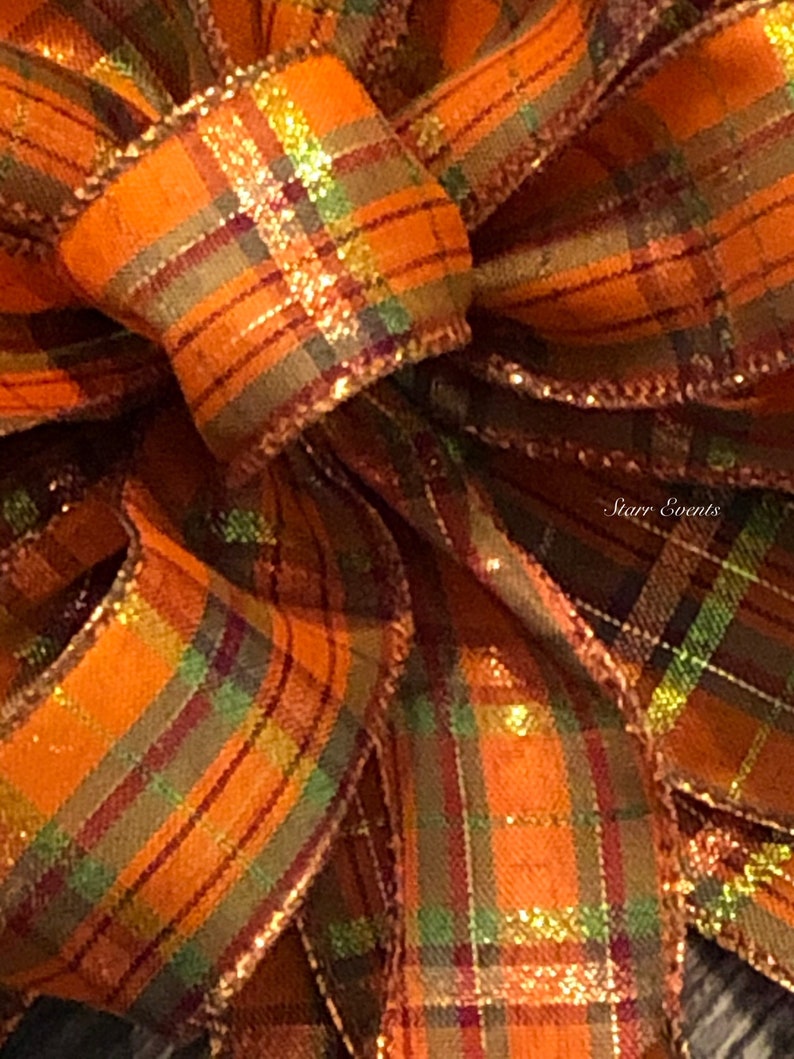 May include: A close-up of a large, orange, green, and gold plaid bow with a shimmery finish. The bow is made of ribbon and is tied in a knot. The bow is perfect for adding a touch of fall decor to any space.