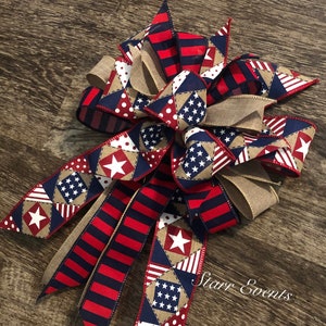 May include: A red, white, and blue patriotic bow with a burlap center. The bow is made of various fabrics with stars and stripes patterns. The bow is tied with a burlap ribbon and has a "Starr Events" tag.