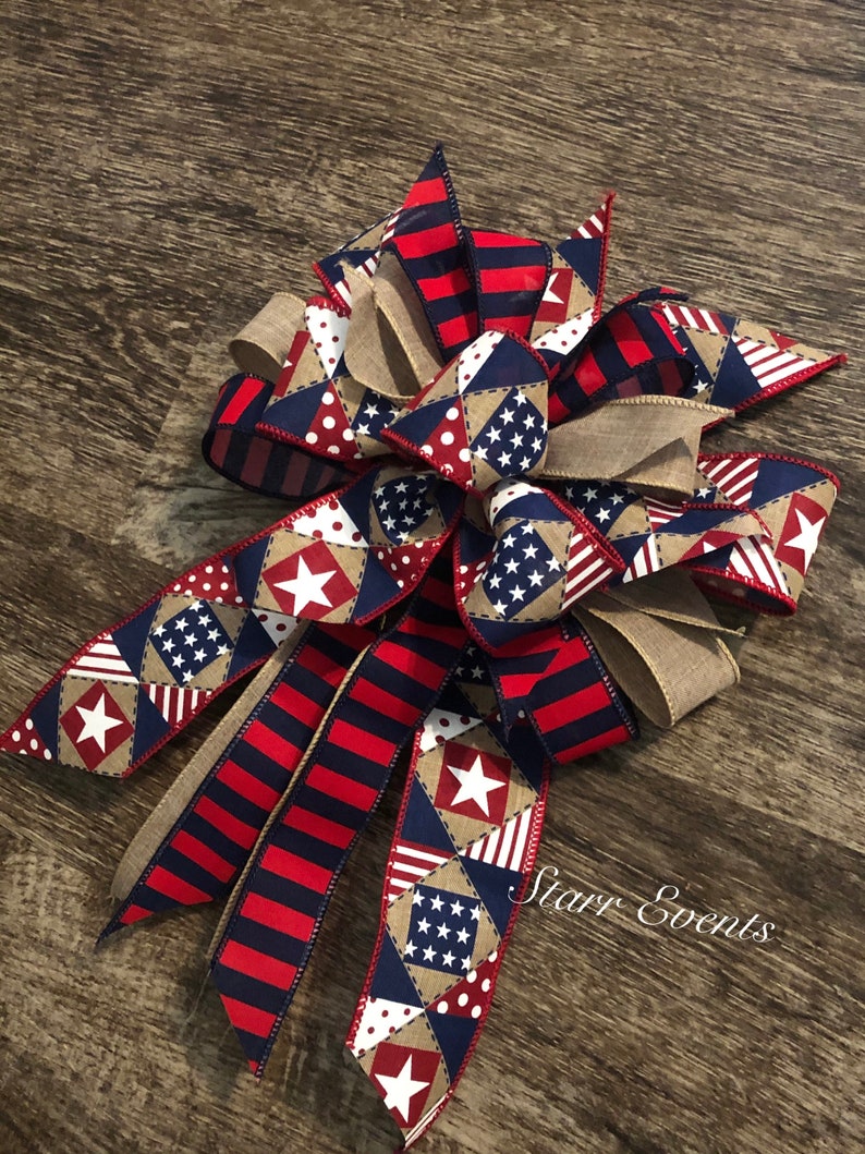 May include: A red, white, and blue patriotic bow with a burlap center. The bow is made of various fabrics with stars and stripes patterns. The bow is tied with a burlap ribbon and has a "Starr Events" tag.