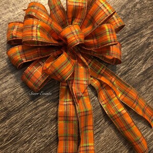 May include: A large orange, green, and gold plaid bow with a wired ribbon tail. The bow is tied with a double loop and has a shimmery finish.