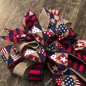 May include: A red, white, and blue patriotic bow with a burlap center. The bow is made of various fabrics with stars, stripes, and polka dots. The bow is tied with a red ribbon and has the text "Starr Events" on the burlap.