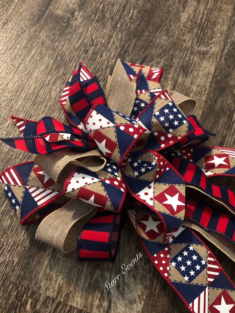 May include: A red, white, and blue patriotic bow with a burlap center. The bow is made of various fabrics with stars, stripes, and polka dots. The bow is tied with a red ribbon and has the text "Starr Events" on the burlap.