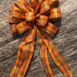 May include: A large orange, green, and gold plaid bow with a long tail. The bow is made of wired ribbon and has a shimmery finish.