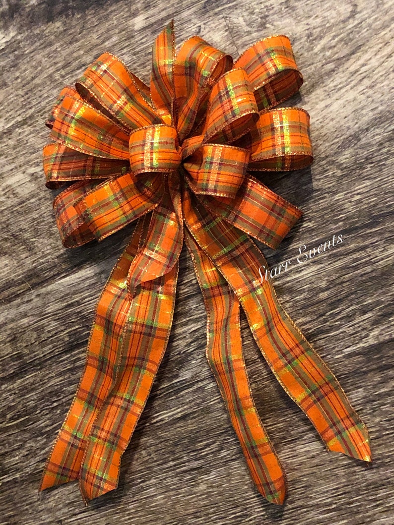 May include: A large orange, green, and gold plaid bow with a long tail. The bow is made of wired ribbon and has a shimmery finish.