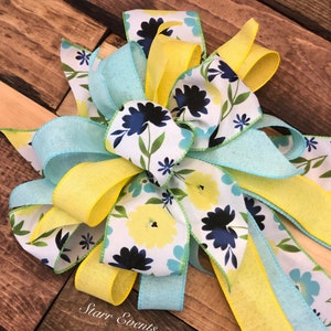 May include: A large, decorative bow made with yellow and blue ribbon. The ribbon has a white floral pattern with blue and yellow flowers. The bow is tied with a double loop and has long tails.