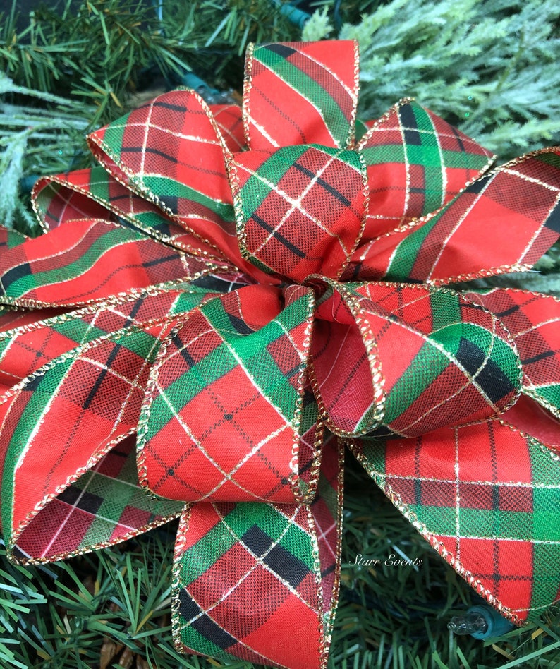 May include: A large, decorative bow with a red, green, black, and gold plaid pattern. The bow is made of fabric and has gold trim along the edges. The bow is likely for Christmas or holiday decorations.