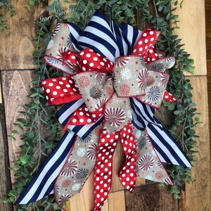 May include: A decorative bow featuring red and white polka dots, navy blue stripes, and a tan fabric with firework and star patterns. The bow is accented with greenery and set against a wooden background.