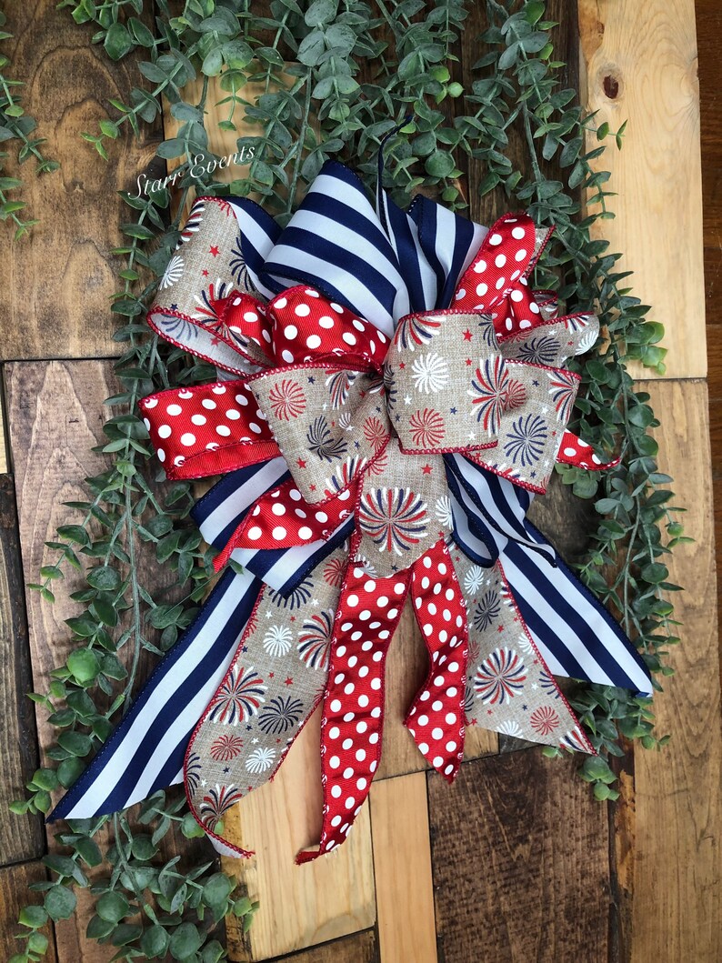 May include: A decorative bow featuring red and white polka dots, navy blue stripes, and a tan fabric with firework and star patterns. The bow is accented with greenery and set against a wooden background.
