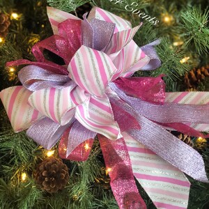 K&ouml;nnte beinhalten: Eine rosa-wei&szlig;e gestreifte Schleife mit silbernen Glitzerakzenten, gebunden an einen gr&uuml;nen Weihnachtsbaumzweig. Die Schleife ist mit einem rosa-silbernen Glitzerband verziert.