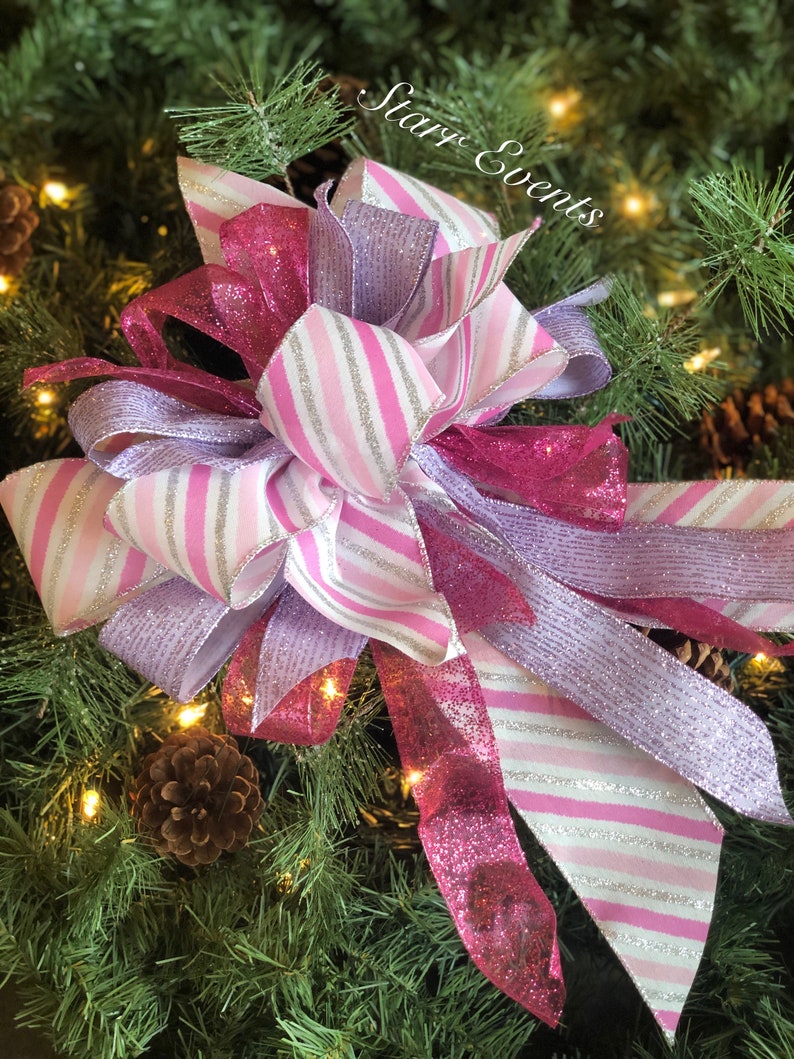 K&ouml;nnte beinhalten: Eine rosa-wei&szlig;e gestreifte Schleife mit silbernen Glitzerakzenten, gebunden an einen gr&uuml;nen Weihnachtsbaumzweig. Die Schleife ist mit einem rosa-silbernen Glitzerband verziert.