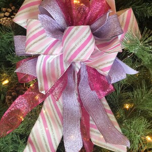 K&ouml;nnte beinhalten: Ein rosa, wei&szlig;er und lavendelfarbener Weihnachtsbaumspitzenbogen mit Glitzer. Die Schleife besteht aus drei verschiedenen B&auml;ndern, einem mit rosa und wei&szlig;en Streifen, einem mit lavendelfarbenen und wei&szlig;en Streifen und einem mit rosa Glitzer. Die Schleife ist mit einem rosa Band gebunden.