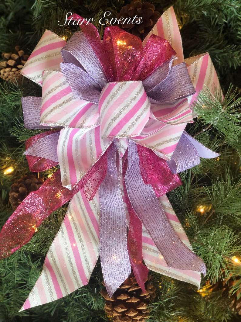 K&ouml;nnte beinhalten: Ein rosa, wei&szlig;er und lavendelfarbener Weihnachtsbaumspitzenbogen mit Glitzer. Die Schleife besteht aus drei verschiedenen B&auml;ndern, einem mit rosa und wei&szlig;en Streifen, einem mit lavendelfarbenen und wei&szlig;en Streifen und einem mit rosa Glitzer. Die Schleife ist mit einem rosa Band gebunden.