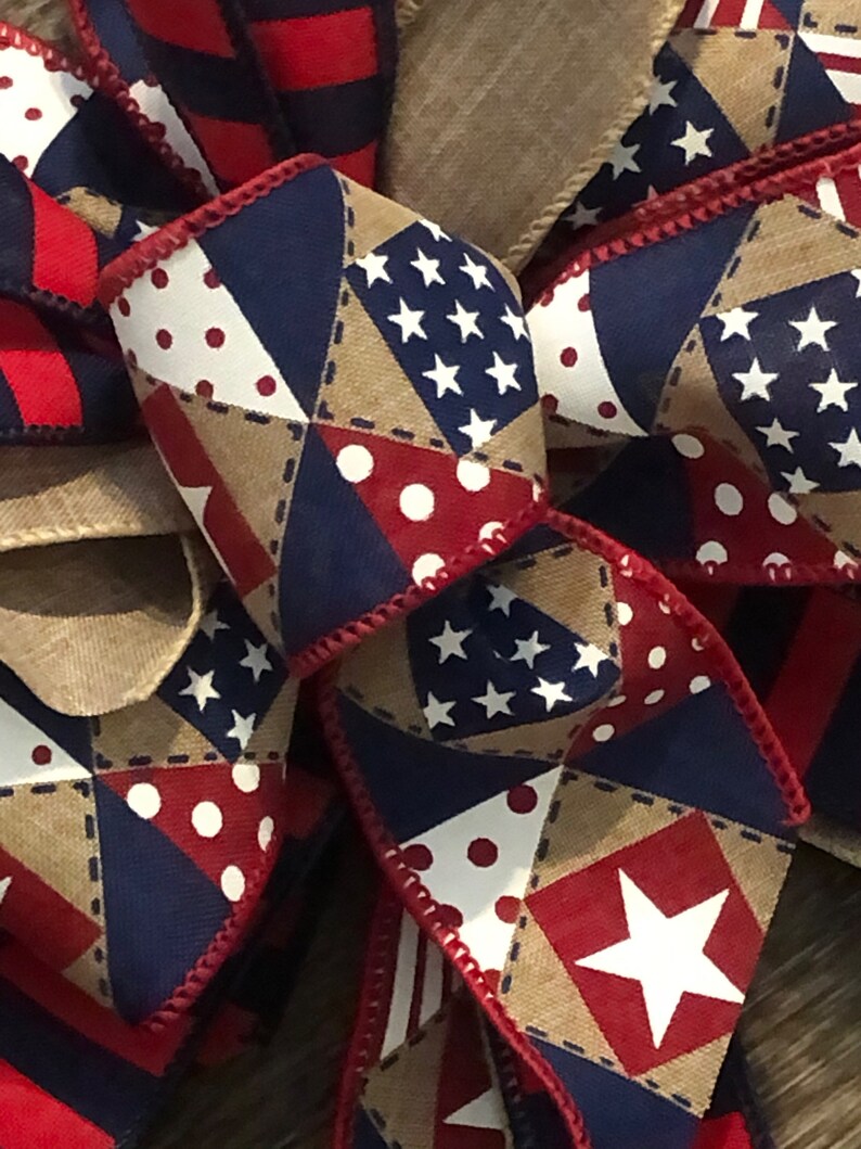 May include: A red, white, and blue patterned ribbon bow with stars, polka dots, and a burlap background. The bow is tied with a red ribbon.
