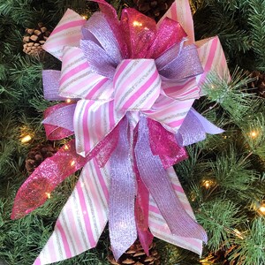K&ouml;nnte beinhalten: Ein rosa-lila Weihnachtsbaumspitzen-Schleife mit silbernen Glitzerstreifen. Die Schleife besteht aus drei verschiedenen B&auml;ndern, einem rosa mit Glitzer, einem lila mit Glitzer und einem wei&szlig;en mit rosa Streifen und silbernem Glitzer. Die Schleife ist oben mit einer Schlaufe gebunden und hat lange Schleifenenden.