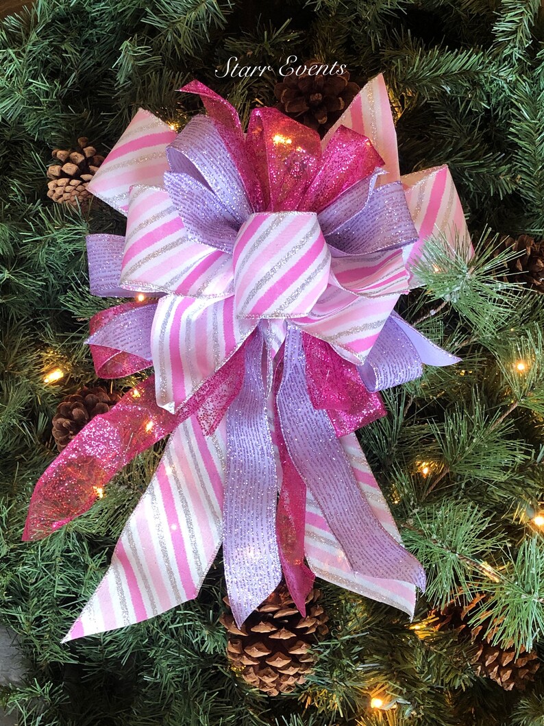 K&ouml;nnte beinhalten: Ein rosa-lila Weihnachtsbaumspitzen-Schleife mit silbernen Glitzerstreifen. Die Schleife besteht aus drei verschiedenen B&auml;ndern, einem rosa mit Glitzer, einem lila mit Glitzer und einem wei&szlig;en mit rosa Streifen und silbernem Glitzer. Die Schleife ist oben mit einer Schlaufe gebunden und hat lange Schleifenenden.